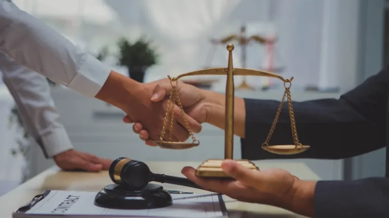 Two individuals shaking hands over a desk with a golden justice scale and a gavel.
