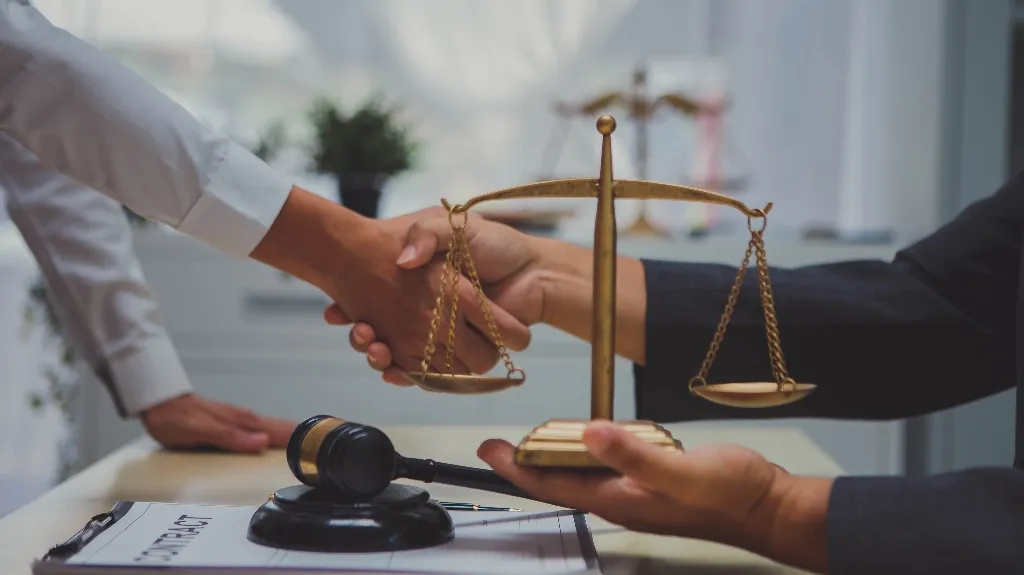 Two individuals shaking hands over a desk with a golden justice scale and a gavel.