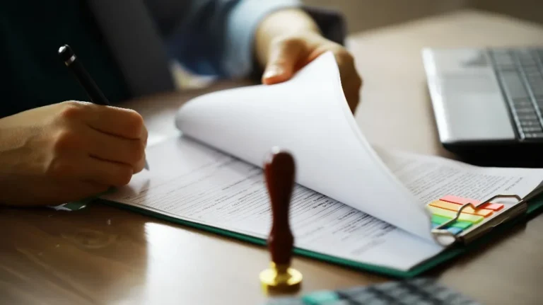 Person signing documents on a clipboard with colorful sticky tabs, laptop, and stamp nearby.