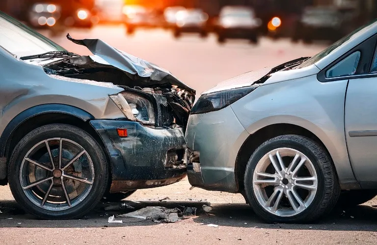Two silver cars with front-end damage after a head-on collision on a road.