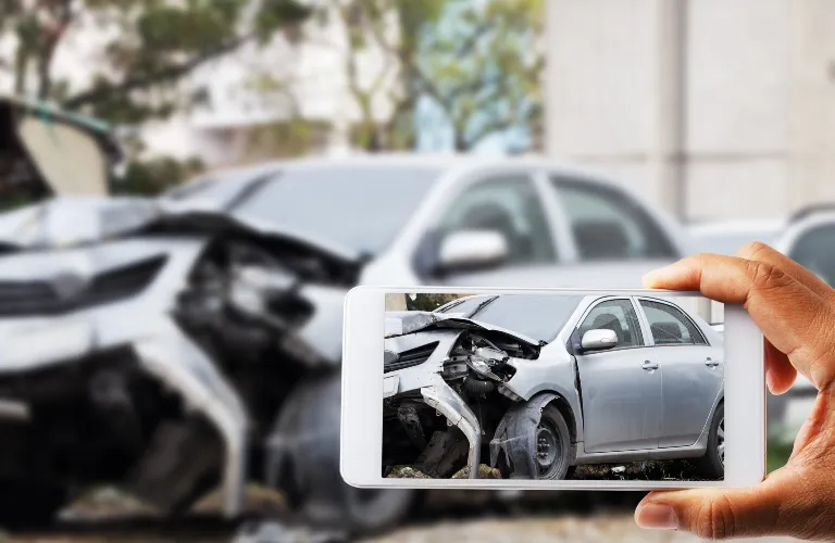Person holding smartphone capturing image of silver car with extensive front-end damage.