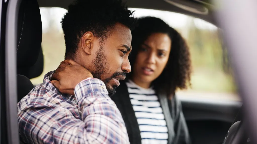 Man in plaid shirt holding neck in discomfort, woman in striped shirt beside him in car.