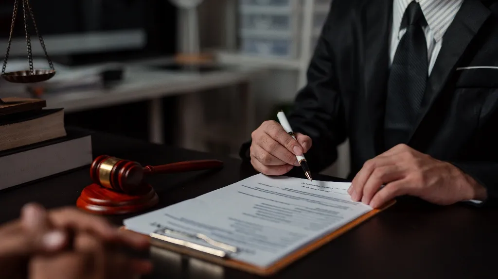 Person in formal suit signing legal document at desk with gavel and scales of justice.