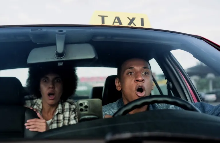 Front view of a male taxi driver and a female passenger inside a red taxi, both showing shocked expressions.