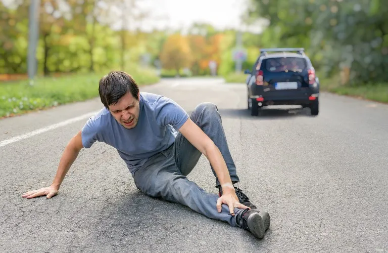 Man in gray clothing sitting on road, holding injured ankle, with a blue car in the background.