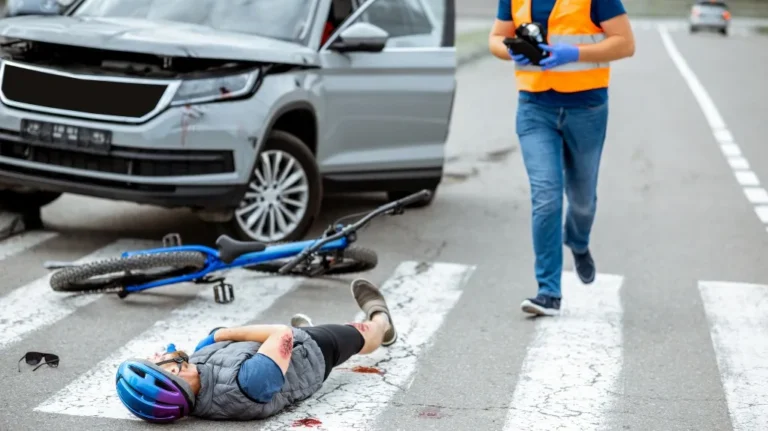 Person wearing a blue helmet lying injured on a crosswalk next to a fallen bicycle and a damaged silver car.