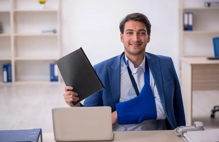 Professional man with arm in blue sling holding black folder, seated at desk with laptop.