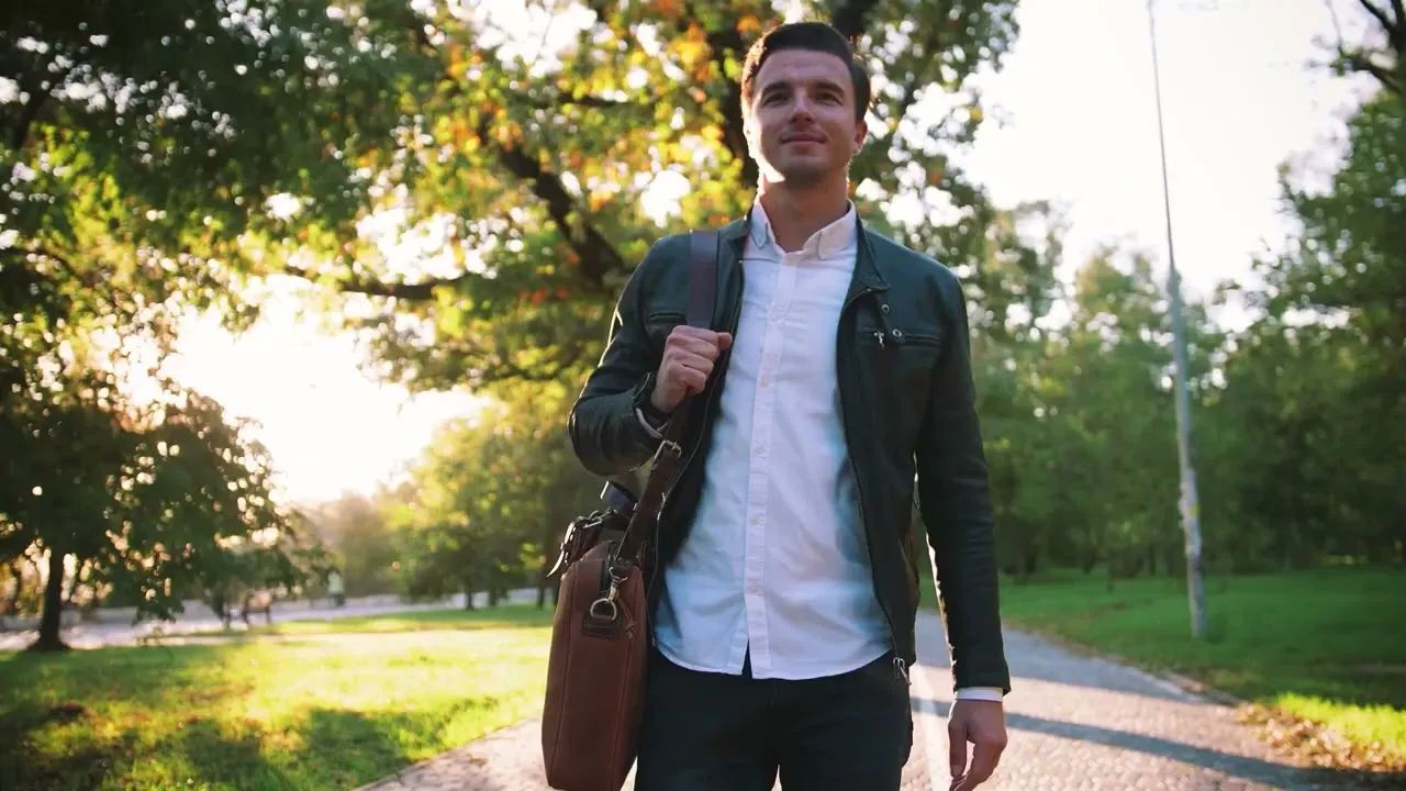 Young man in black jacket and white shirt carrying brown bag walking in sunlit park.