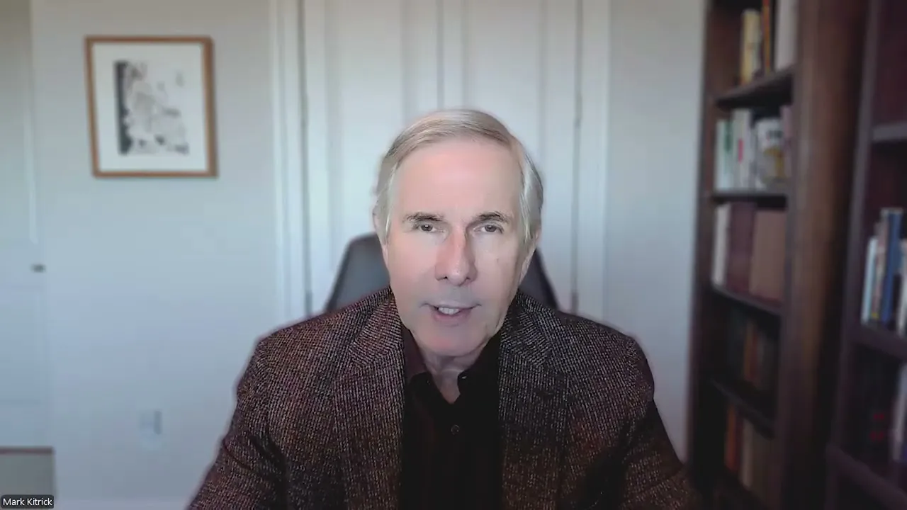 Middle-aged man with gray hair wearing a dark blazer, seated in an office with bookshelves and framed artwork.