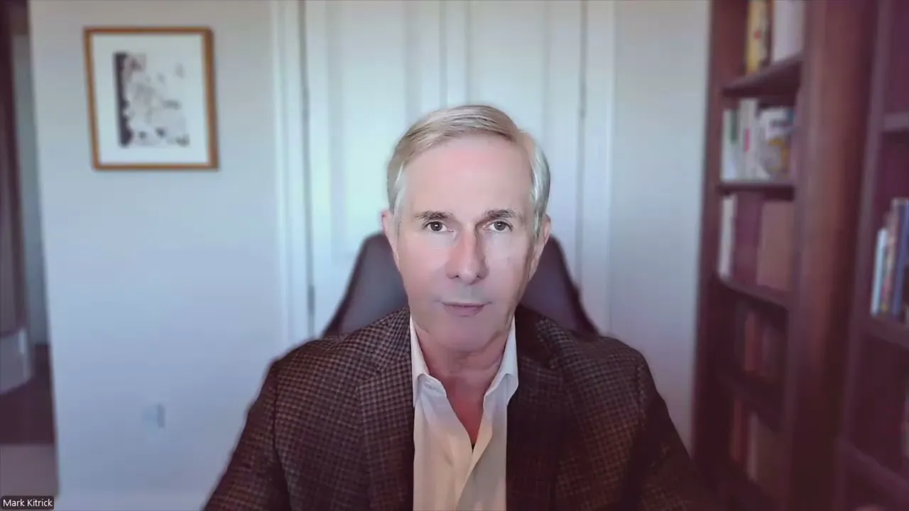 Middle-aged man with gray hair wearing a checkered blazer and white shirt, seated in a home office with bookshelves and framed artwork.