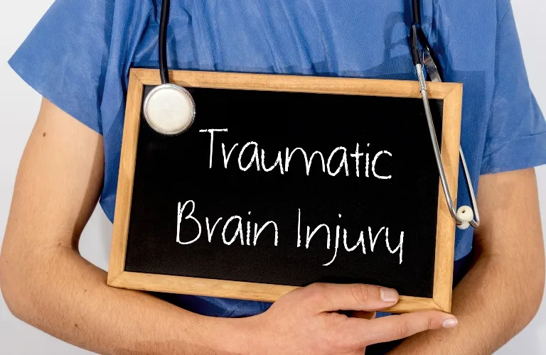 Person in blue medical scrubs holding a chalkboard with "Traumatic Brain Injury" text and a stethoscope.