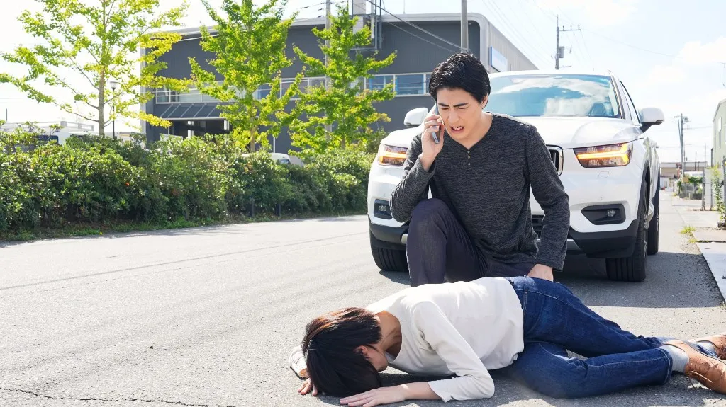 Man making a phone call beside a woman lying unconscious on the pavement near a white car.