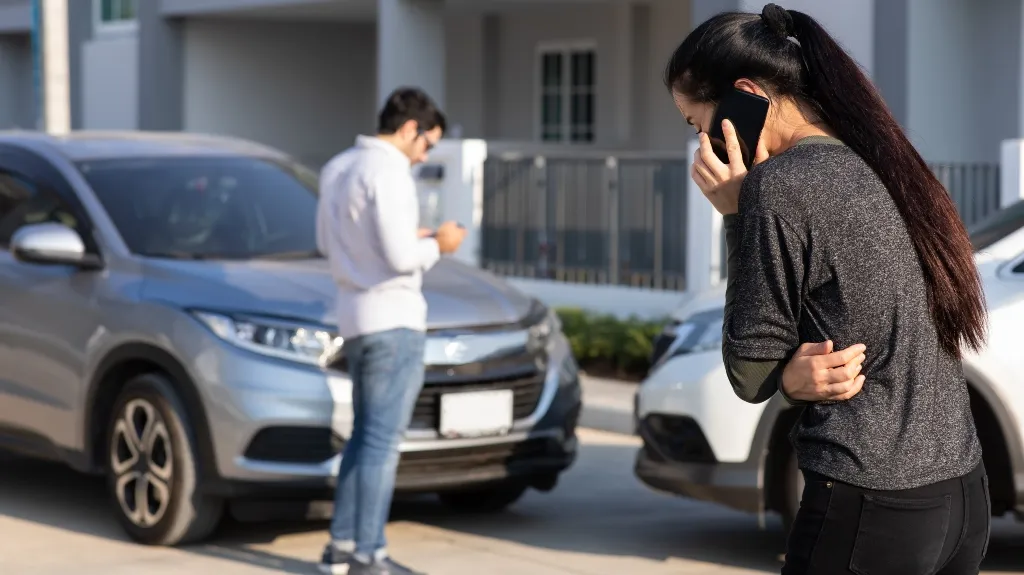 Two individuals near a silver and white car, one woman on phone, one man checking his device.