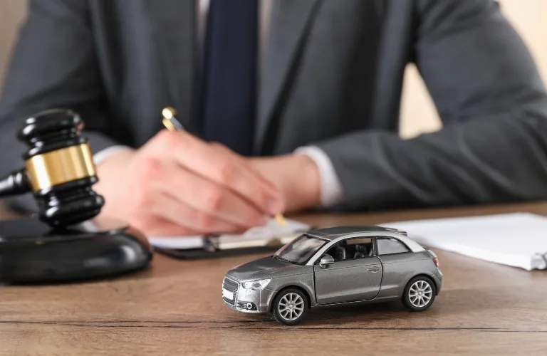 Gray toy car on wooden desk with gavel and person in suit writing in background.