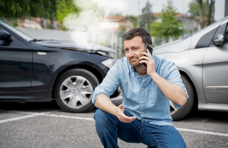 Man in blue shirt making a phone call beside two damaged cars in a parking lot.