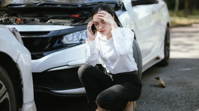 Woman in white shirt and black pants kneeling beside damaged white car, speaking on phone.