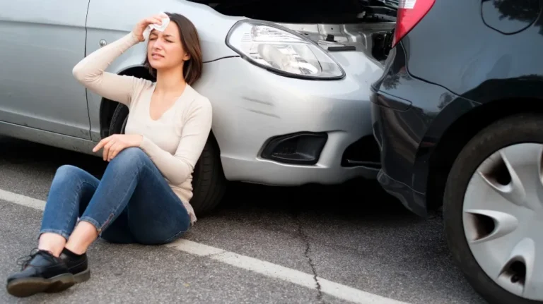 Woman sitting on pavement beside two cars involved in a rear-end collision.