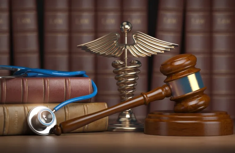 Wooden judge's gavel, medical caduceus symbol, stethoscope, and legal books on a desk.