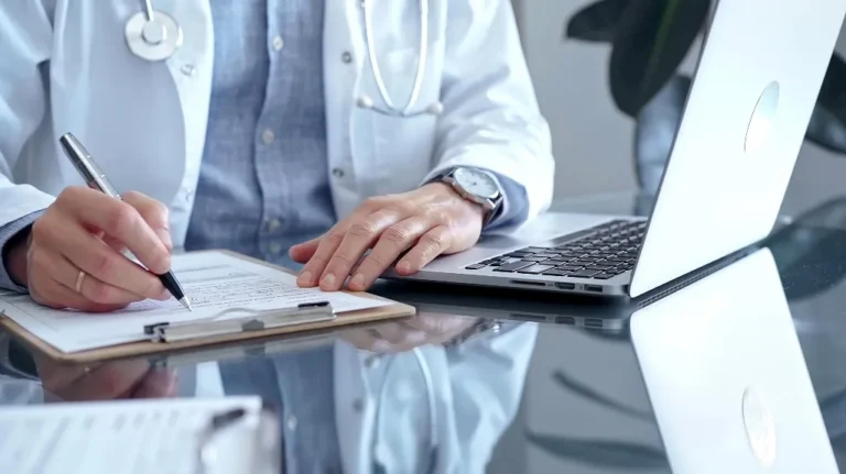 Medical professional in white coat writing on clipboard beside open laptop on glass desk.