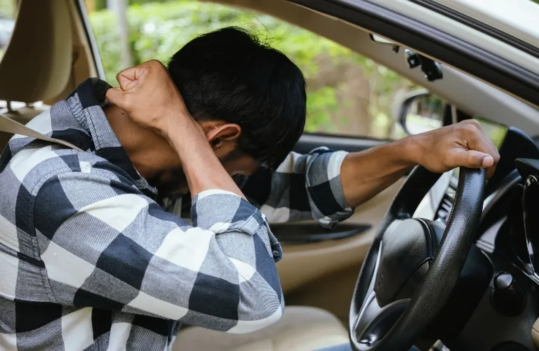 Man in black and white checkered shirt sitting in car, leaning forward with head down and hands on neck.